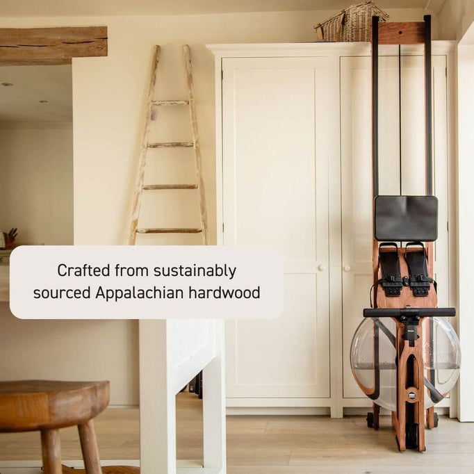 A rowing machine with a wooden frame and black handlebars stands in a bright, modern room with cream cabinets, a wooden stool, and a ladder. A text overlay reads 'Crafted from sustainably sourced Appalachian hardwood'.