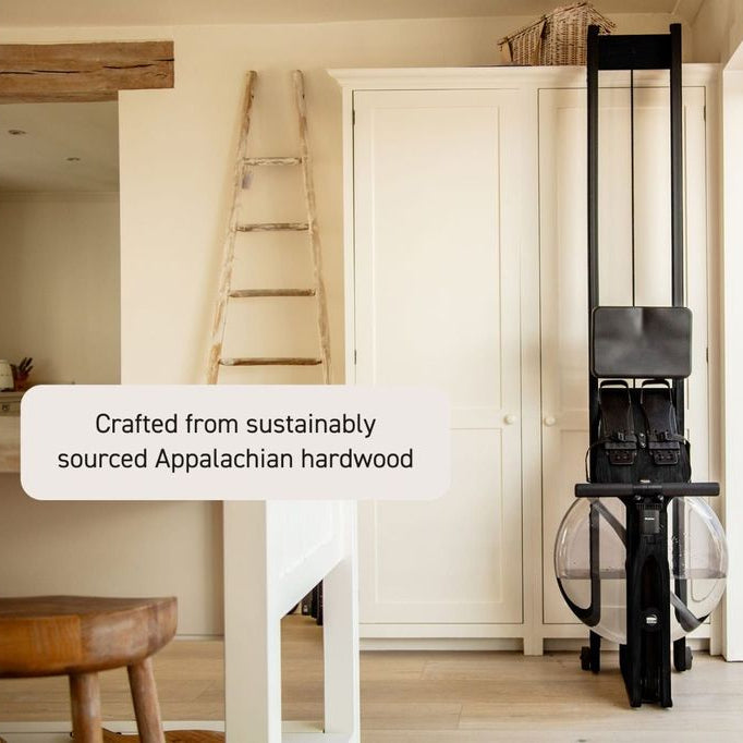 A Pilates reformer machine with a black frame and clear weight-bearing discs, set in a bright, modern indoor space with a wooden floor and white walls. A text overlay on the left reads 'Crafted from sustainably sourced Appalachian hardwood'.