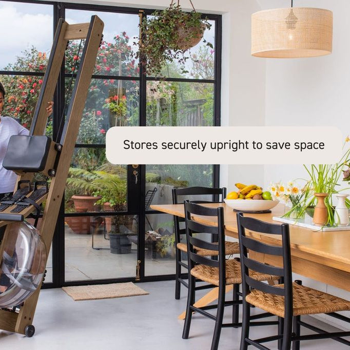 A wooden rowing machine in a bright modern home gym with large black-framed glass doors, a dining table, chairs, and a hanging pendant light. A text bubble reads 'Stores securely upright to save space'.