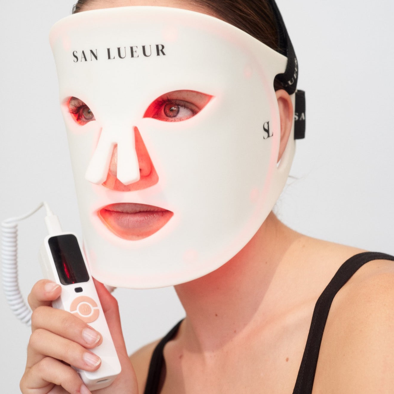 Person wearing a LED face mask with 'SAN LUEUR' branding, holding a device.