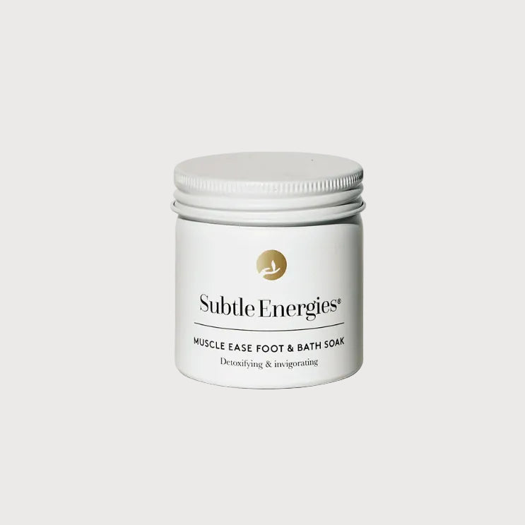 A white jar of Subtle Energies Muscle Ease Foot & Bath Soap with a silver lid, set on a light beige textured surface against a white background.
