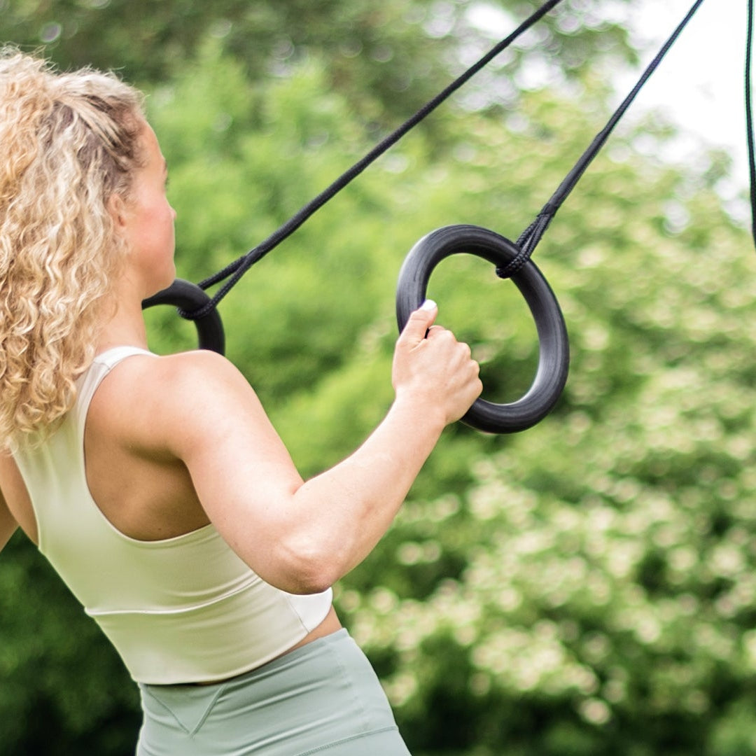 A woman with curly blonde hair exercises on black gymnastic rings outdoors in a park with green trees in the background.