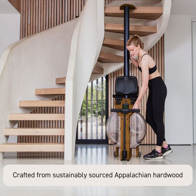 A woman in black activewear stands next to a rowing machine placed in a modern interior with a spiral staircase and wood slat wall. Text at the bottom reads, 'Crafted from sustainably sourced Appalachian hardwood.