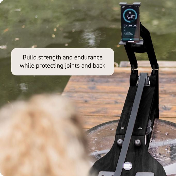 A black pet back carrier support stand with a smartphone mounted on top, shown on a wooden dock with water in the background. A white text bubble reads 'Build strength and endurance while protecting joints and back'.