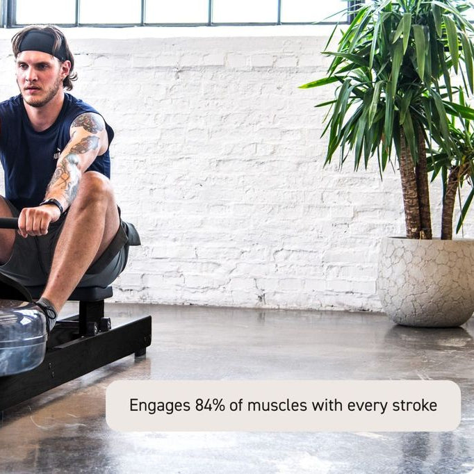 A man wearing a black headband and athletic clothing is using a black rowing machine in a bright indoor space with a white brick wall, concrete floor, and a potted plant. A text overlay at the bottom right reads: 'Engages 84% of muscles with every stroke.