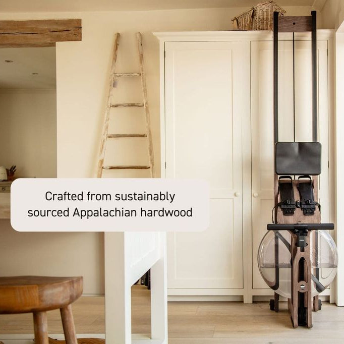 A rowing machine with a wooden frame and black handlebars stands in a bright, modern room with cream cabinets, a wooden stool, and a ladder. A text overlay reads 'Crafted from sustainably sourced Appalachian hardwood'.