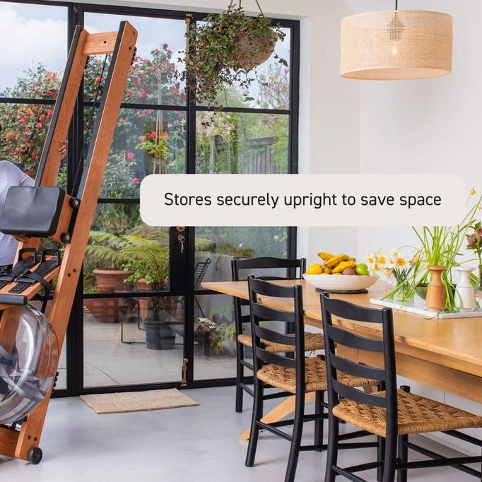 A woman stands next to a wooden rowing machine in a bright modern room with large windows, a dining table, and chairs. A text bubble says 'Stores securely upright to save space'.