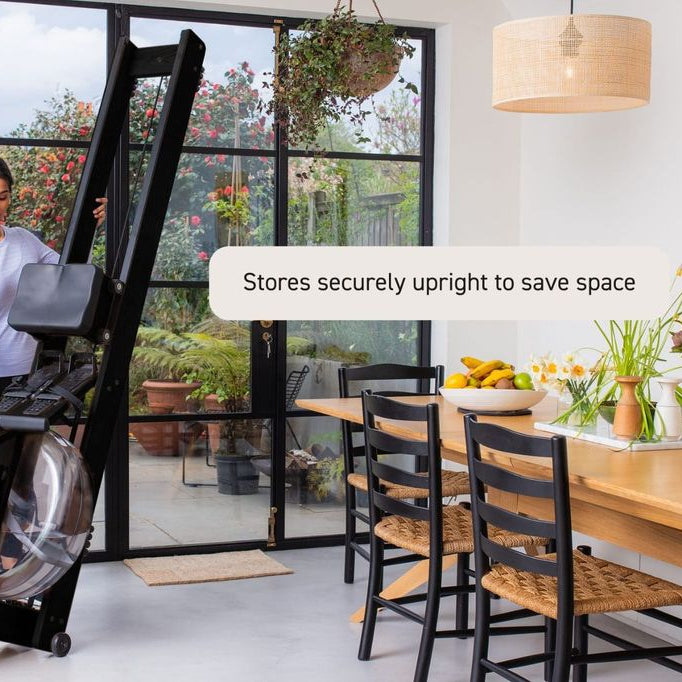 A woman in a home office setting demonstrates a black rowing machine, showing how it folds flat and stores upright. A text overlay reads 'Stores securely upright to save space'.