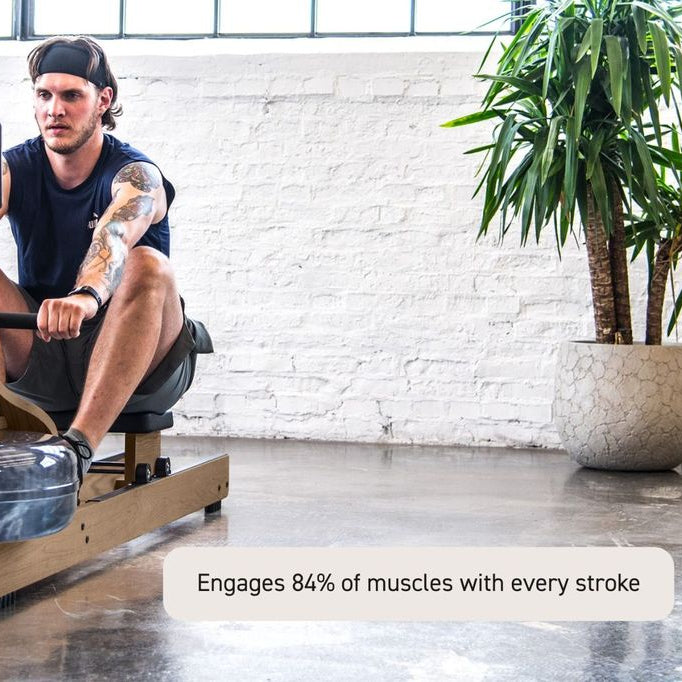A man with tattoos sits on a rowing machine in a bright indoor gym space with a white brick wall, concrete floor, and a large potted plant. A text overlay at the bottom reads, 'Engages 84% of muscles with every stroke'.